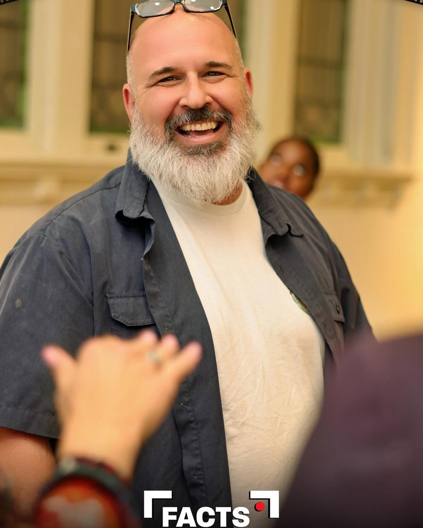 Smiling man with glasses on head, beard, wearing a dark shirt and light t-shirt, in a room with windows and people.