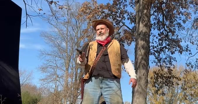Man outdoors wearing a hat, vest, and scarf, standing among trees with blue sky behind him.