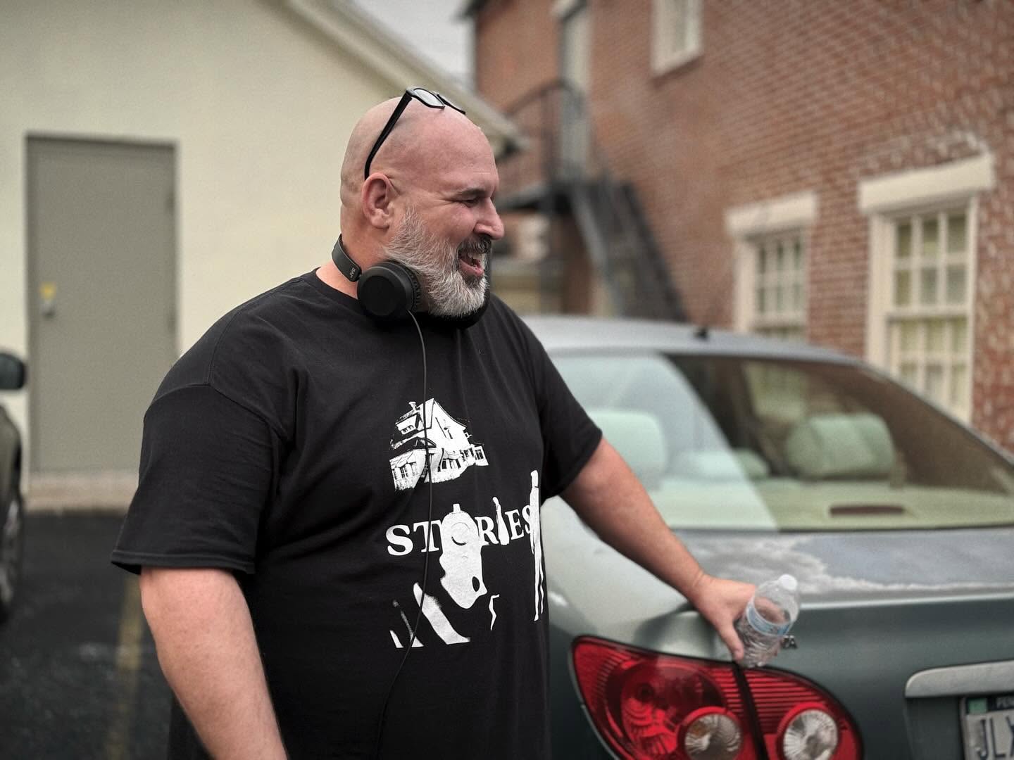 Man with glasses on head, headphones around neck, smiling, standing outdoors next to a car, holding a water bottle.