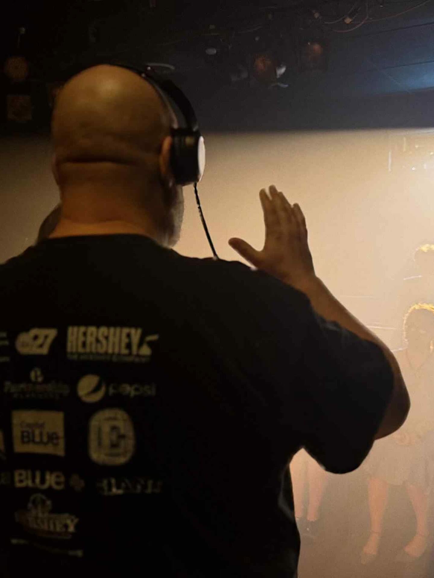 Person with headphones facing a wall, raising their hand, wearing a black shirt with logos on the back, in a dimly lit room.