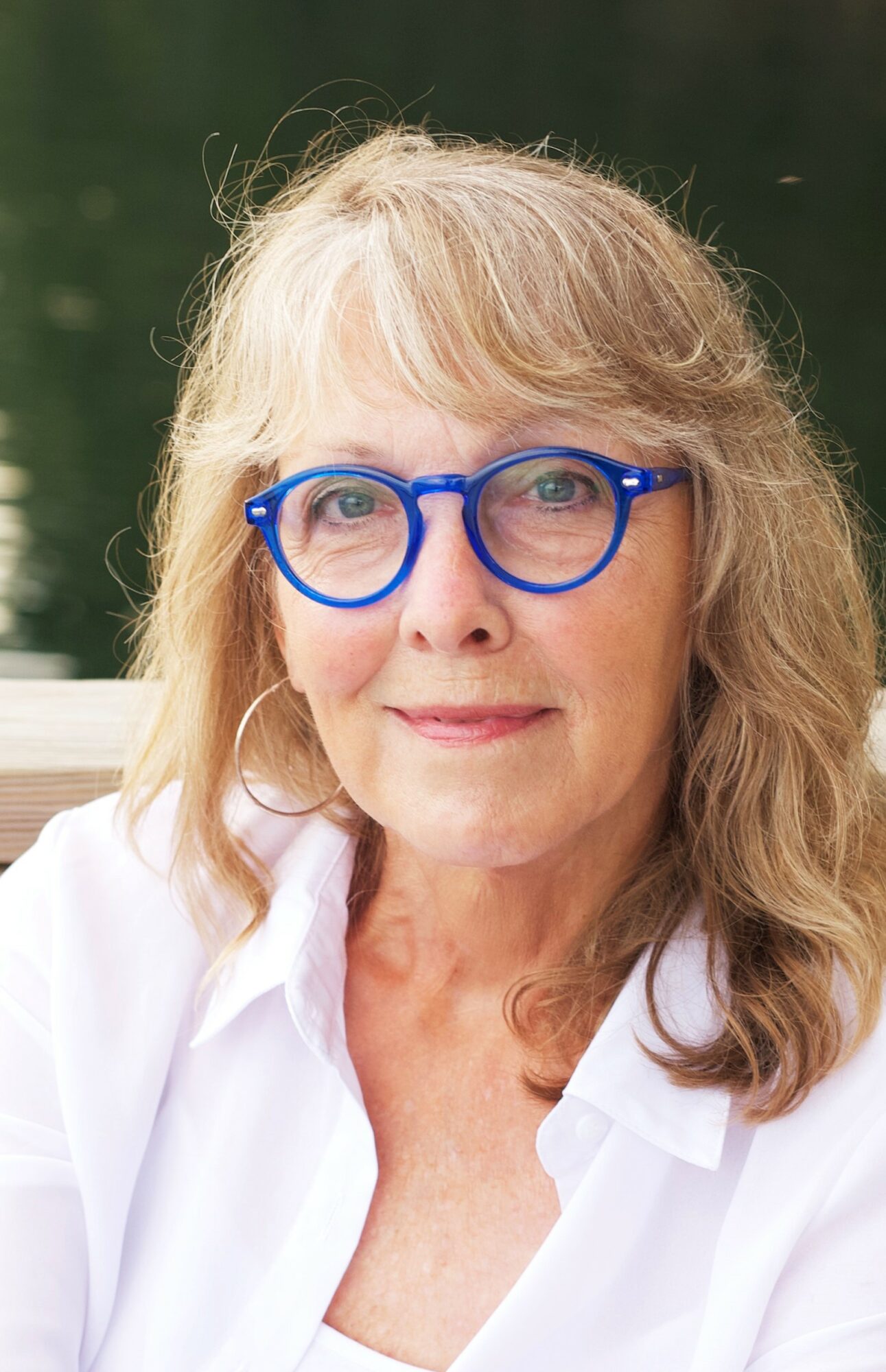 Woman with blonde hair, blue glasses, wearing a white shirt outdoors, smiling softly.