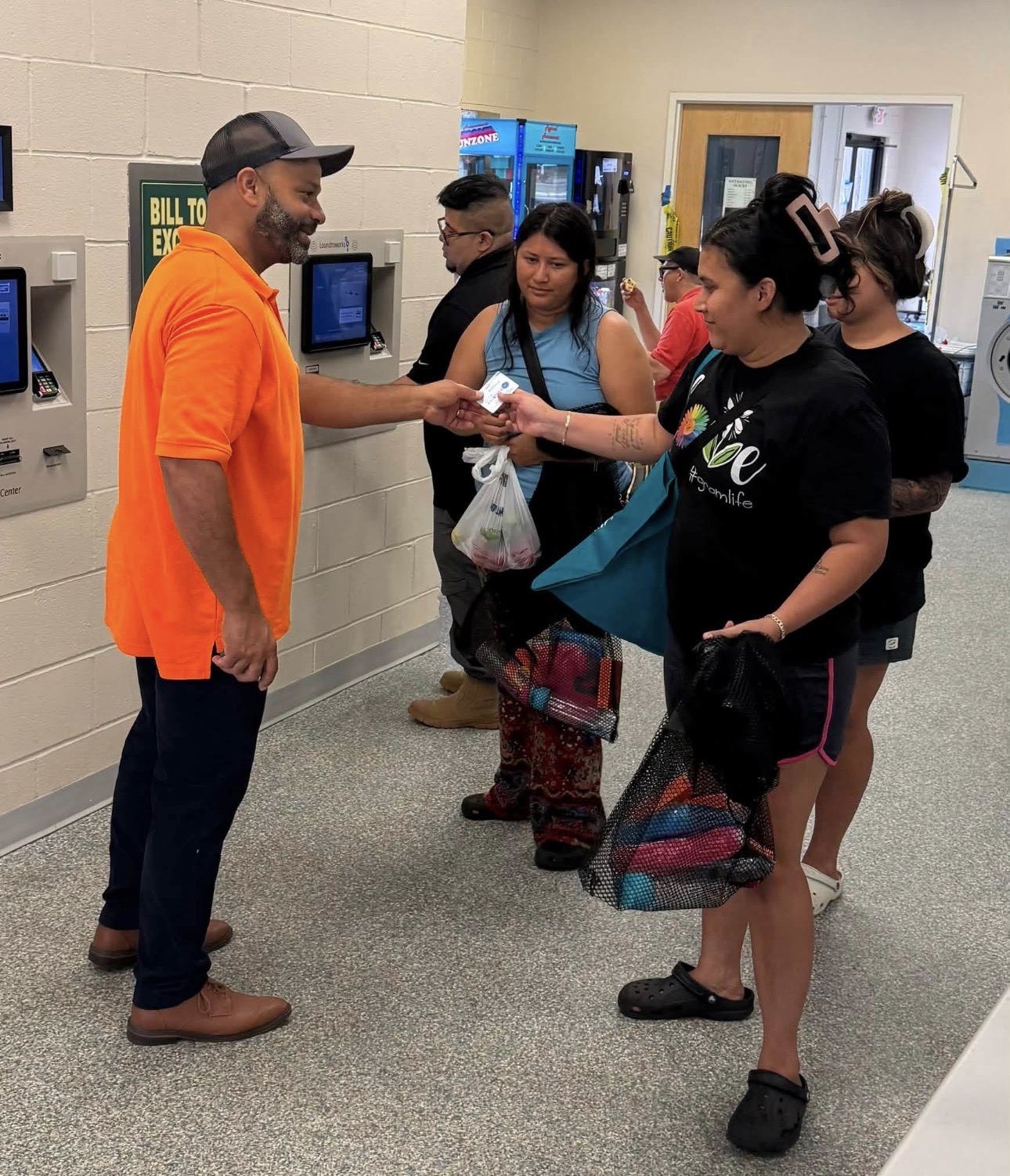 Person in orange shirt hands item to person in black shirt in a hallway with other people and vending machines.