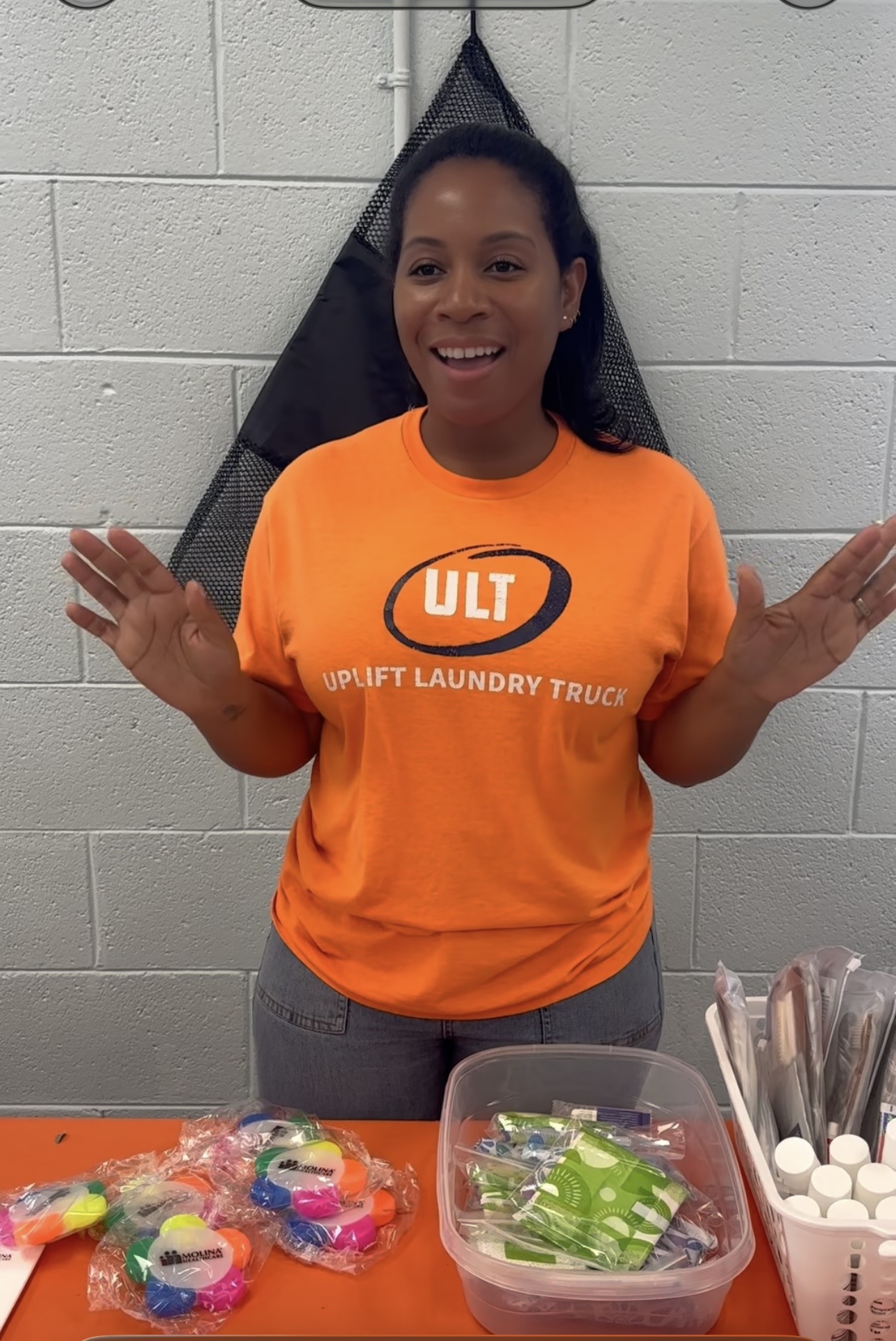 Smiling woman in orange T-shirt with ULIT logo, standing against a white wall, hands raised, in a laundry room.