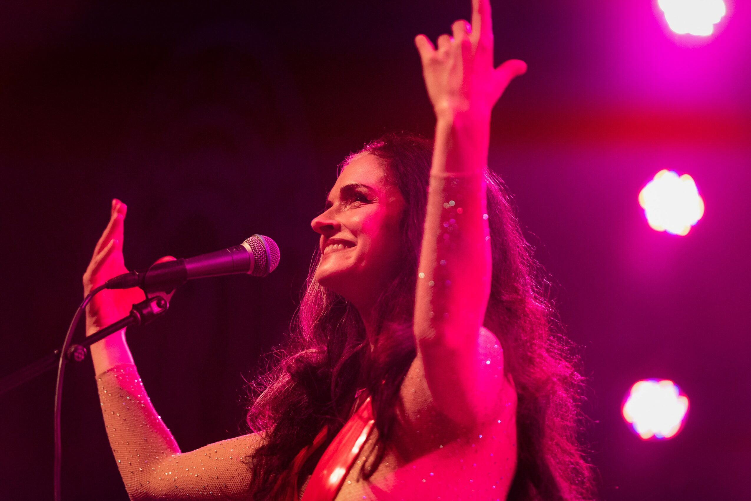 Woman smiling and raising her hands on stage with pink and purple lighting, microphone in front of her.