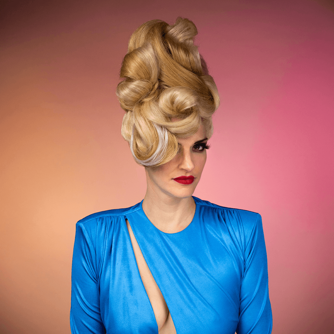 Woman with elaborate blonde hairstyle and red lipstick wearing a shiny blue dress against a pink background.