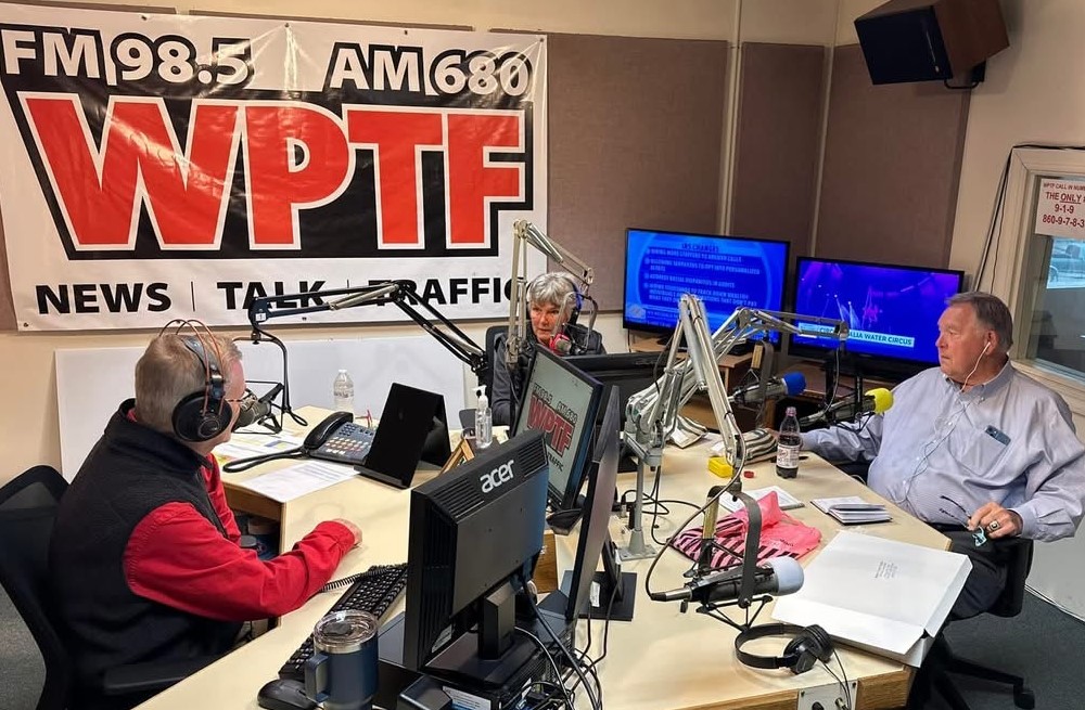 Three people in a radio studio with microphones, computers, and a large WPTF sign on the wall.