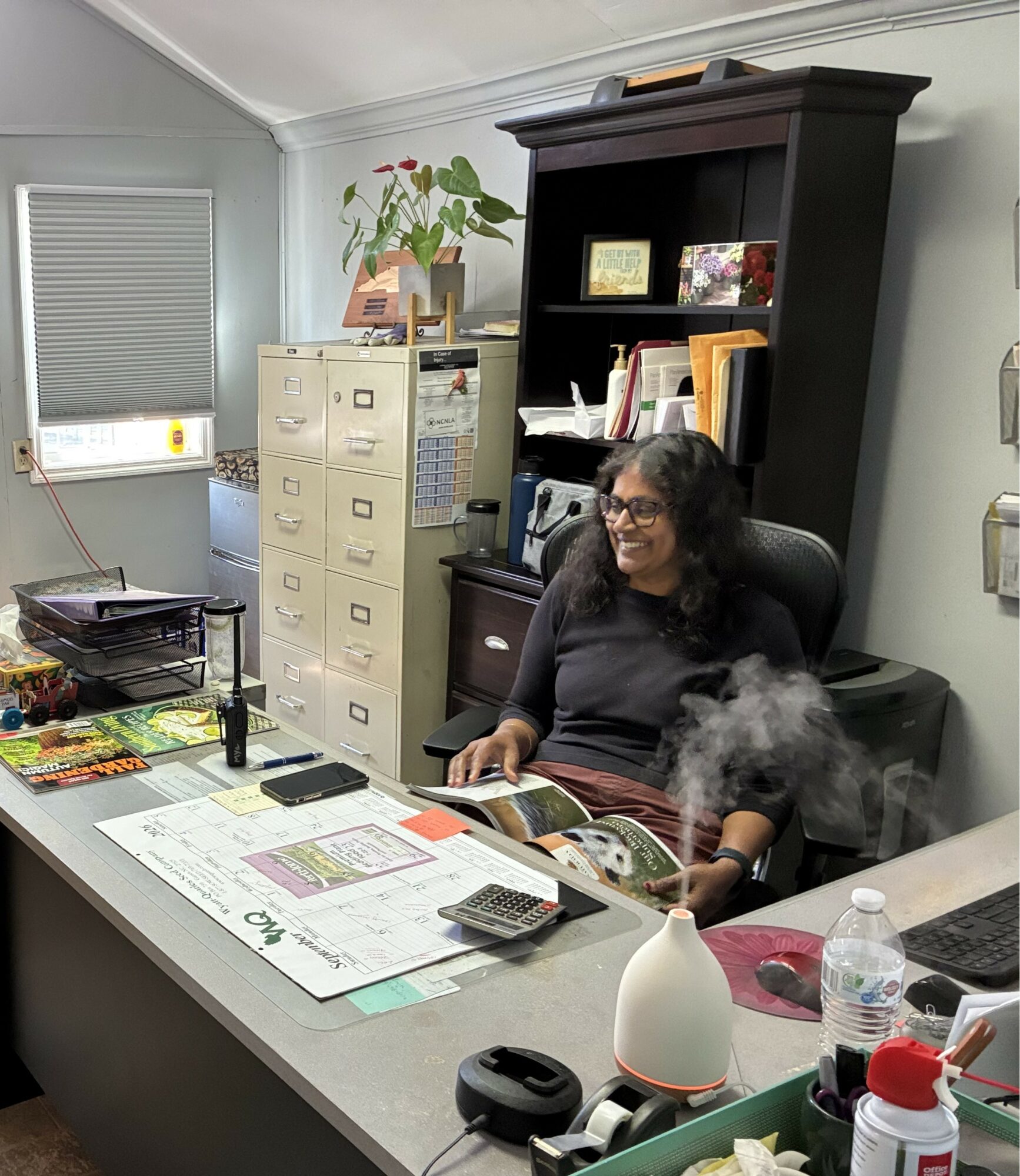 Woman sitting at desk in office, smiling, with books, papers, and office supplies on desk, bookshelf behind her, and a filing cabinet nearby.