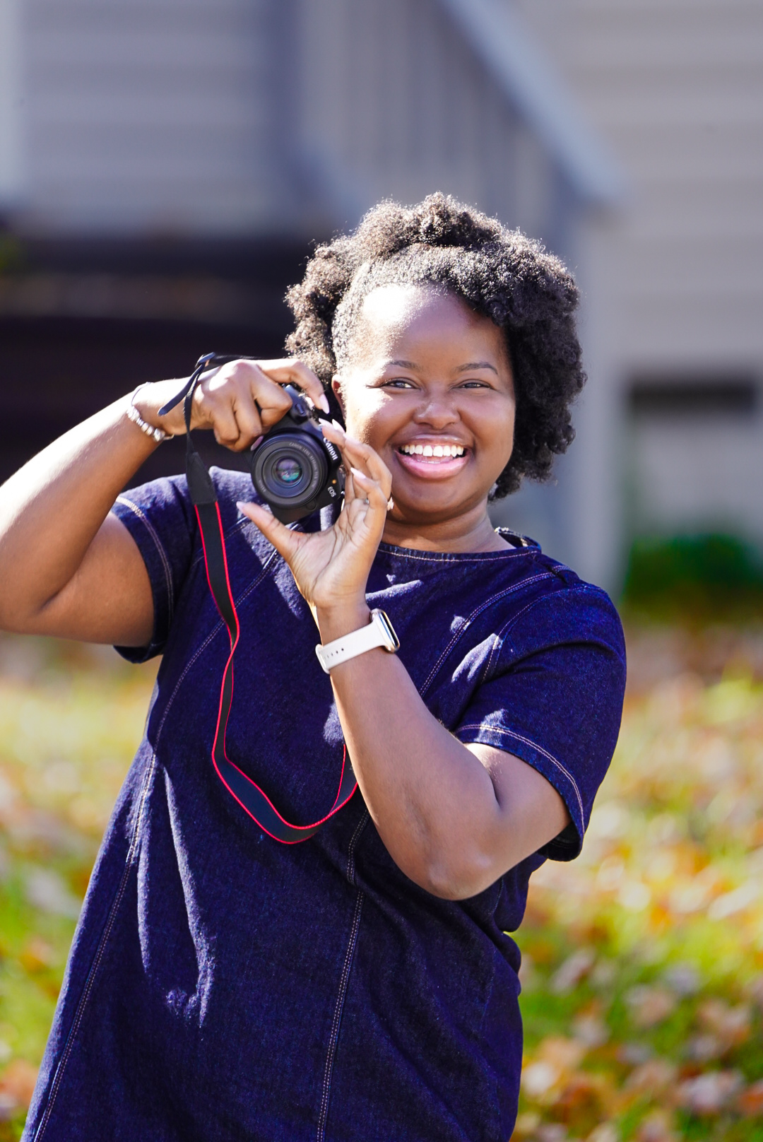 Smiling woman holding a camera outdoors, wearing a navy blue shirt, with a house and greenery in the background.