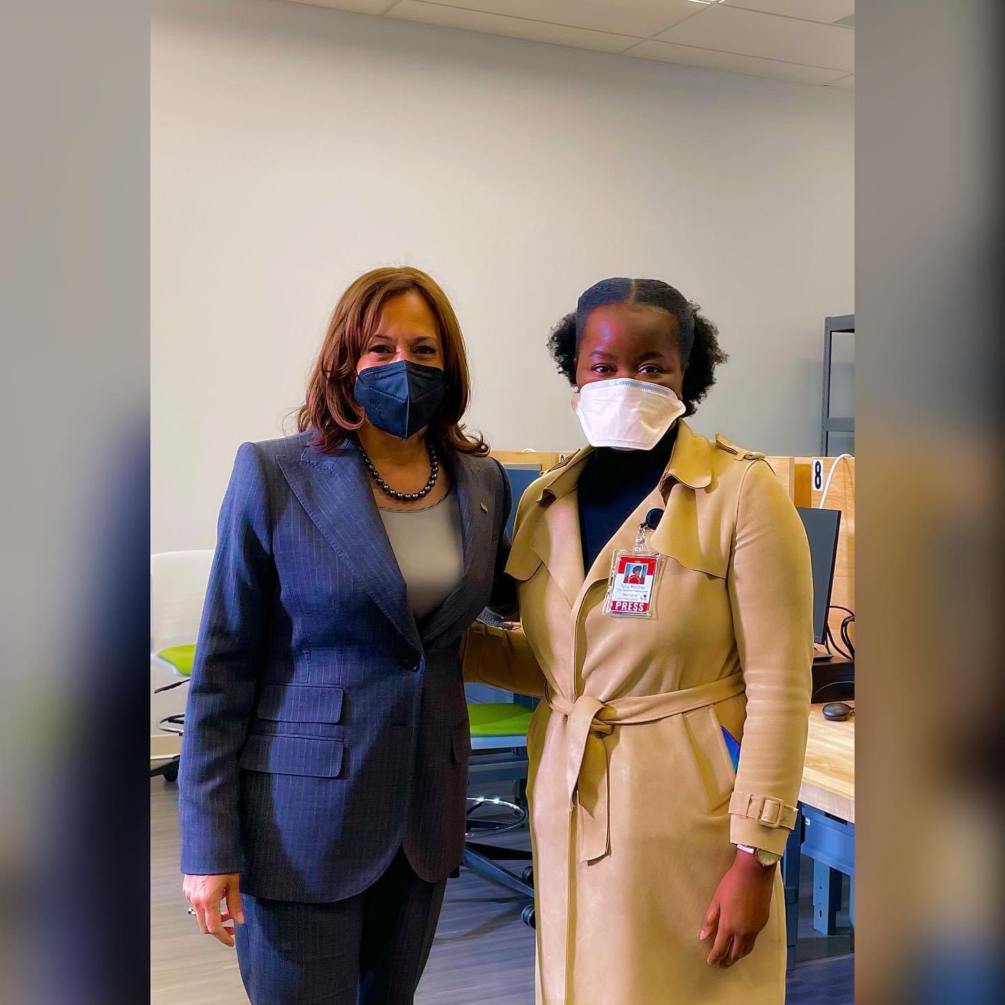 Two women wearing masks standing indoors, one in a blue suit and the other in a beige coat, in front of a plain wall.