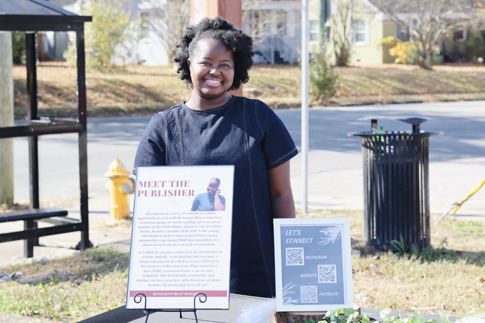 Smiling person standing outdoors near informational signs, with houses and trees in the background.