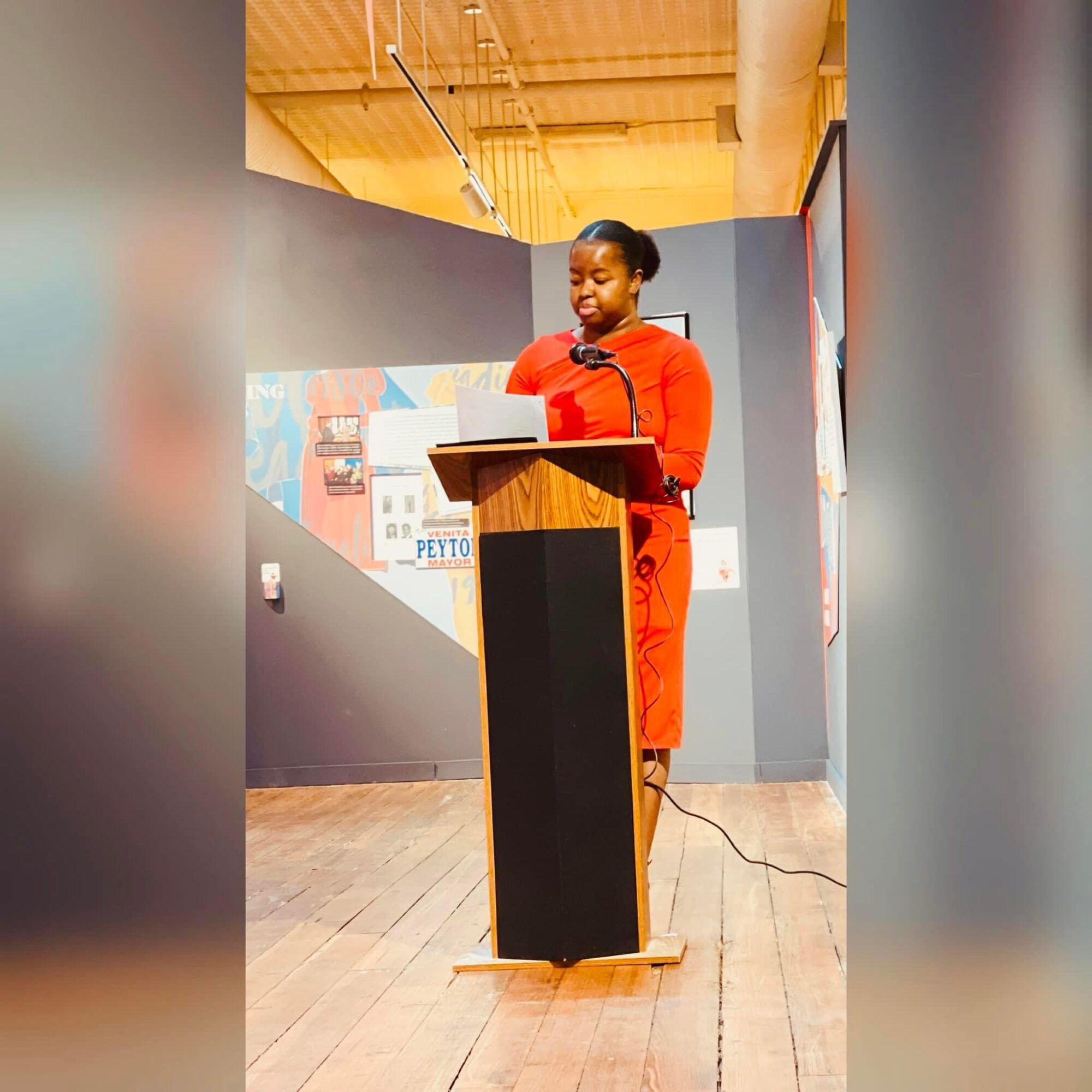 Woman in red dress standing at a podium, speaking or presenting in a room with wooden floor and gray walls.