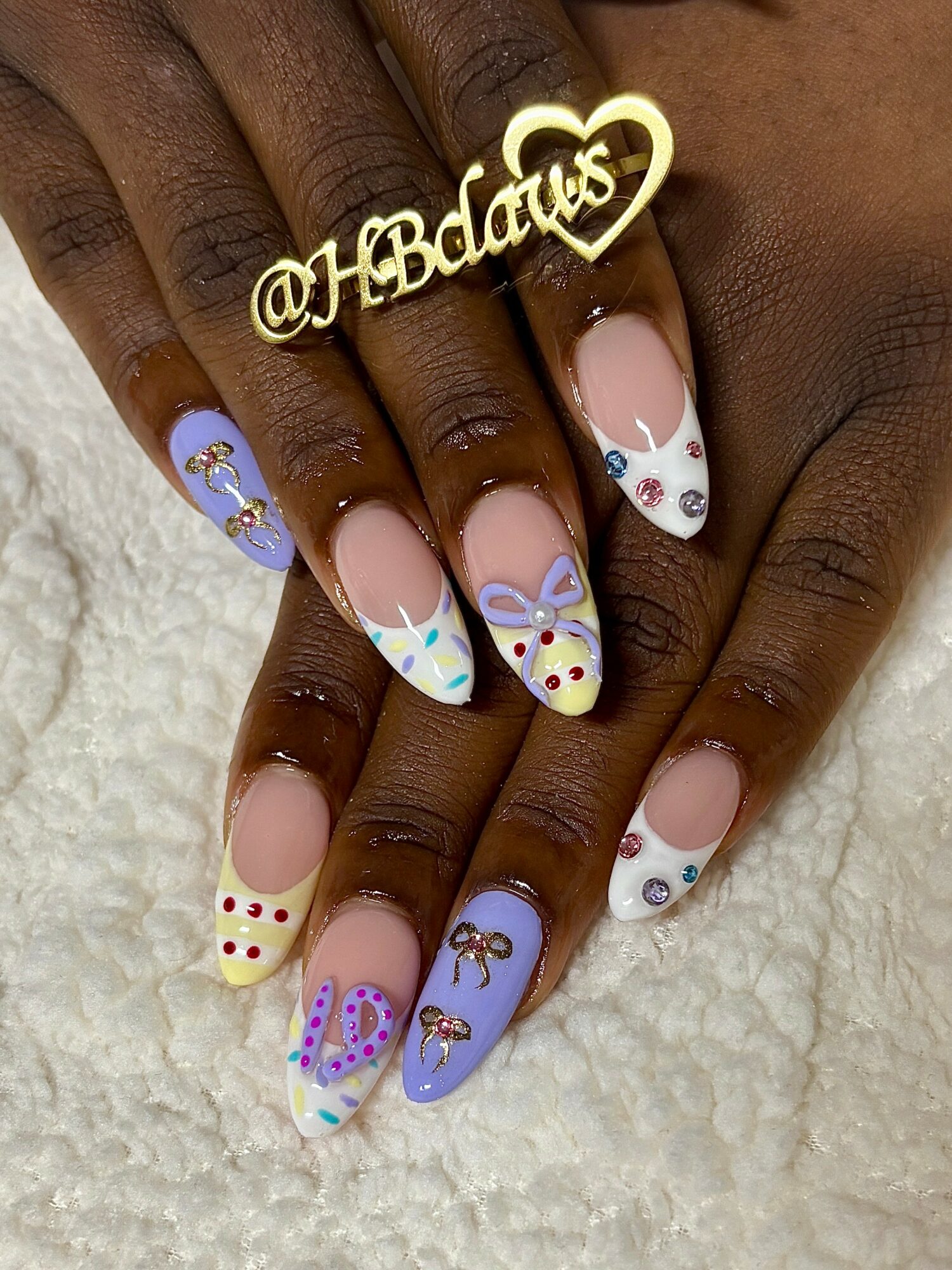 Hands with decorated nails featuring pastel colors, hearts, dots, and bows, holding a gold heart-shaped sign with '@HBabies'.
