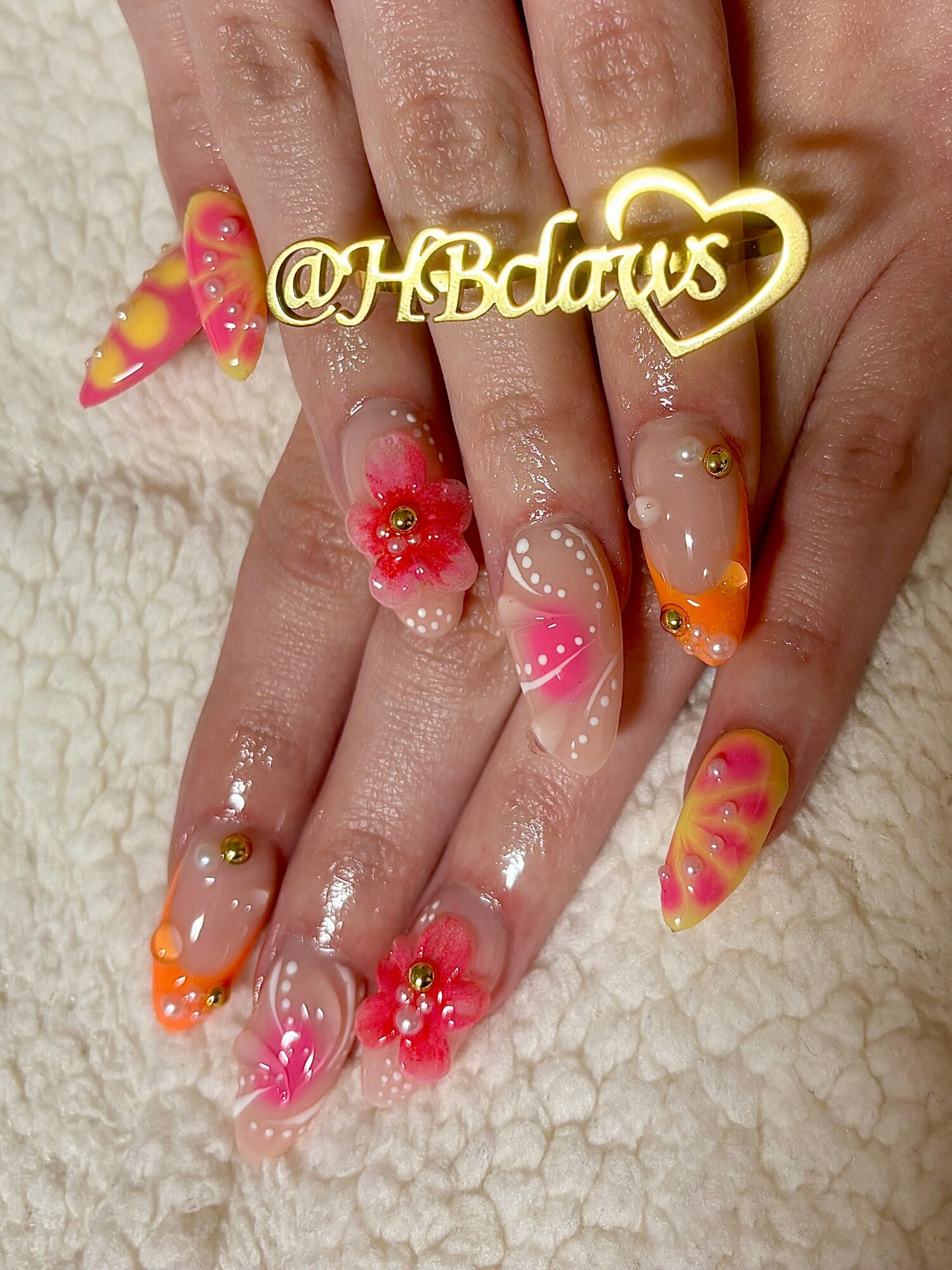 Hands with colorful, decorated nails featuring pink, orange, and yellow designs, flowers, and rhinestones, resting on a soft surface.