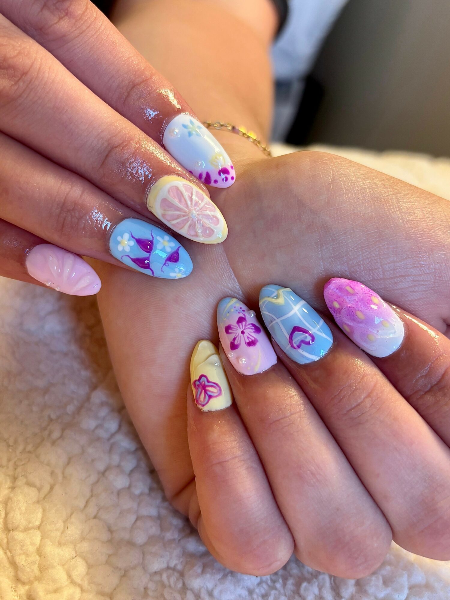 Two hands with colorful, decorated nails resting on a soft surface, displaying various nail art designs with flowers and patterns.