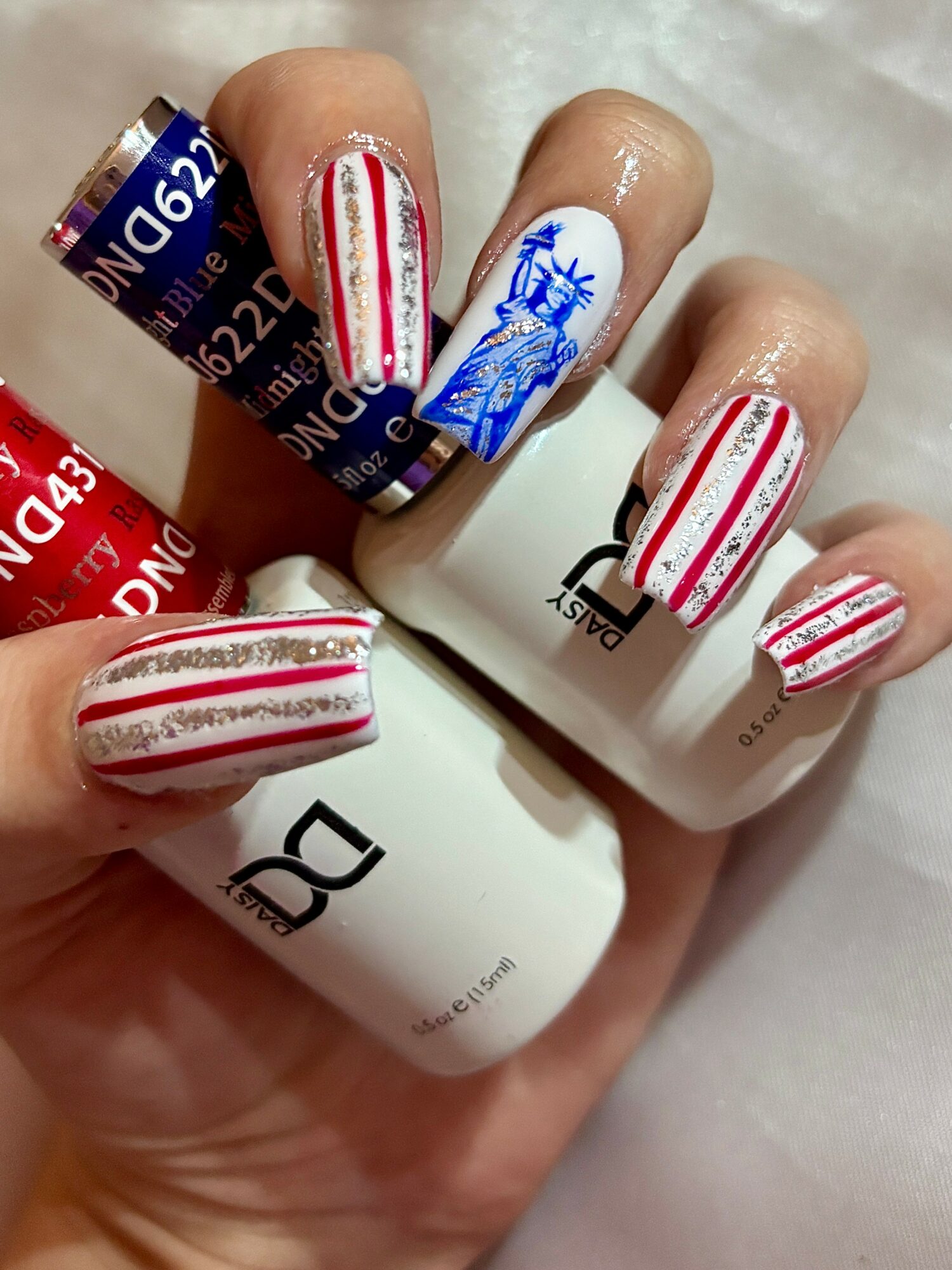 Hand with nails painted in red and white stripes, one nail features a blue design, holding nail polish bottles.