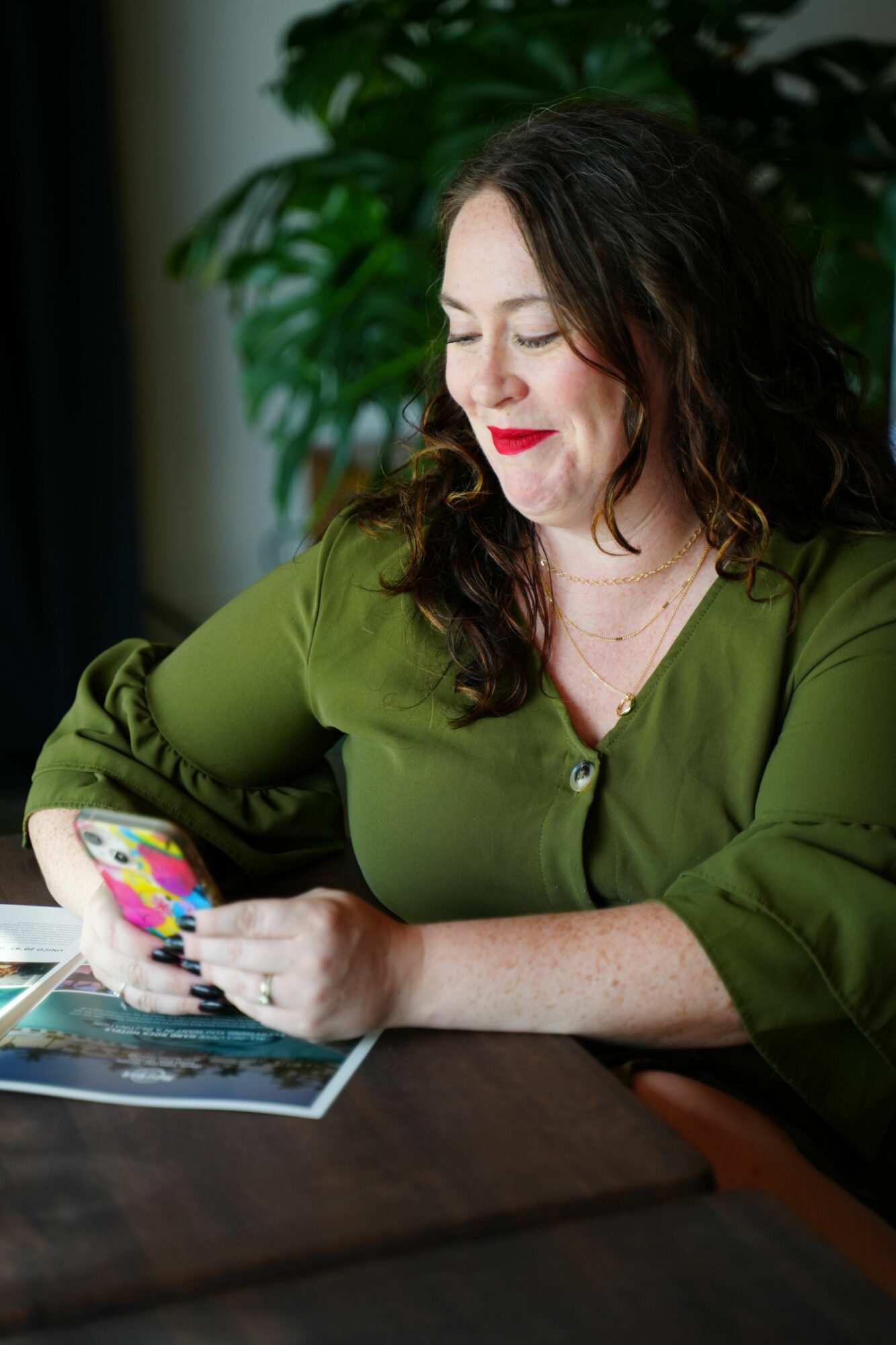 Woman with dark hair and red lipstick reads a colorful book at a table, with green plants in the background.