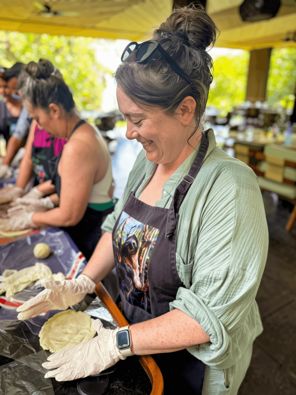 Woman smiling, wearing apron and gloves, participating in outdoor cooking activity with others in background.
