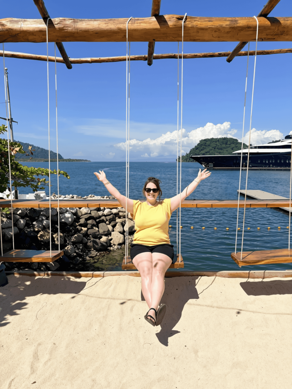 Woman sitting on a swing with arms outstretched, by water with boats and islands in background, sunny day.