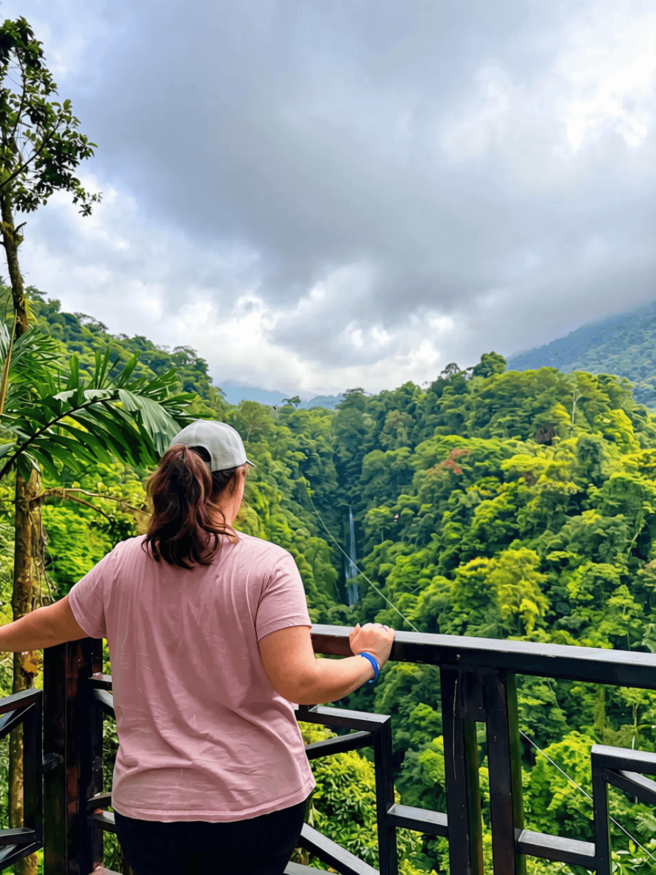 Person with long hair, wearing a pink shirt and gray cap, standing on a balcony overlooking a lush green forested landscape under cloudy sky.
