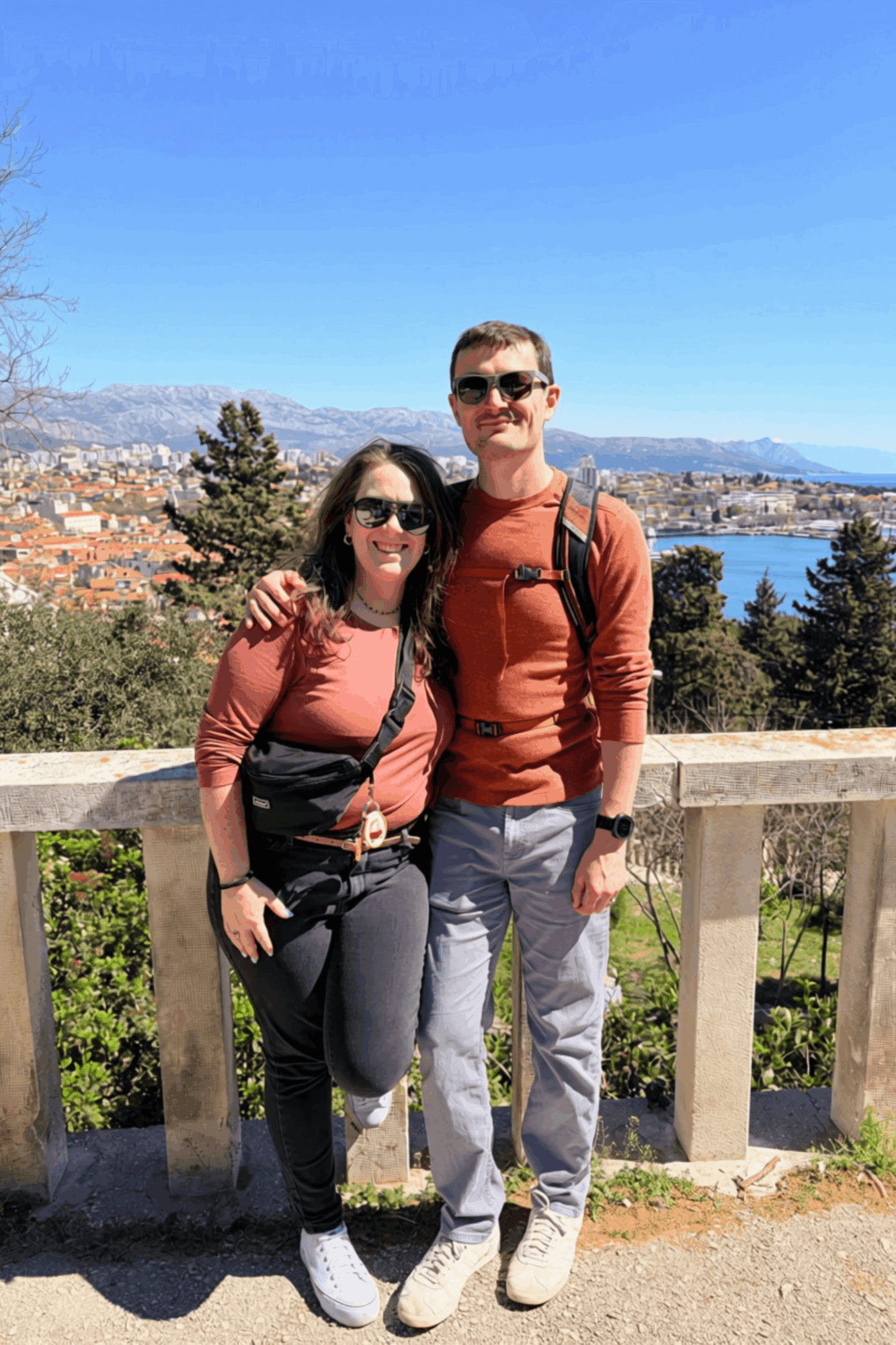 A man and woman stand outdoors on a bridge with a scenic view of water, trees, and mountains behind them.