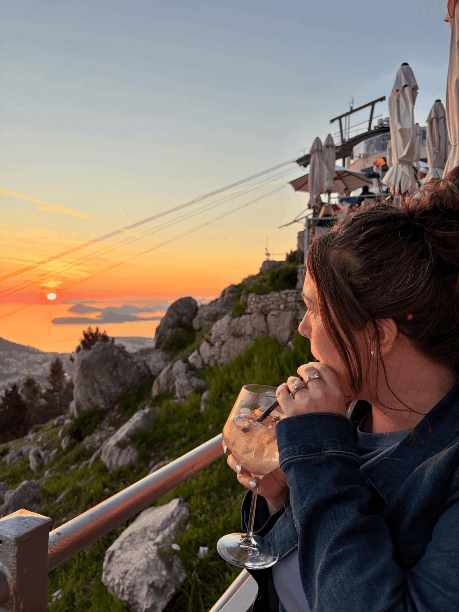 Person with dark hair in a ponytail holding a glass of drink, overlooking a sunset over rocky coastline with umbrellas and chairs.