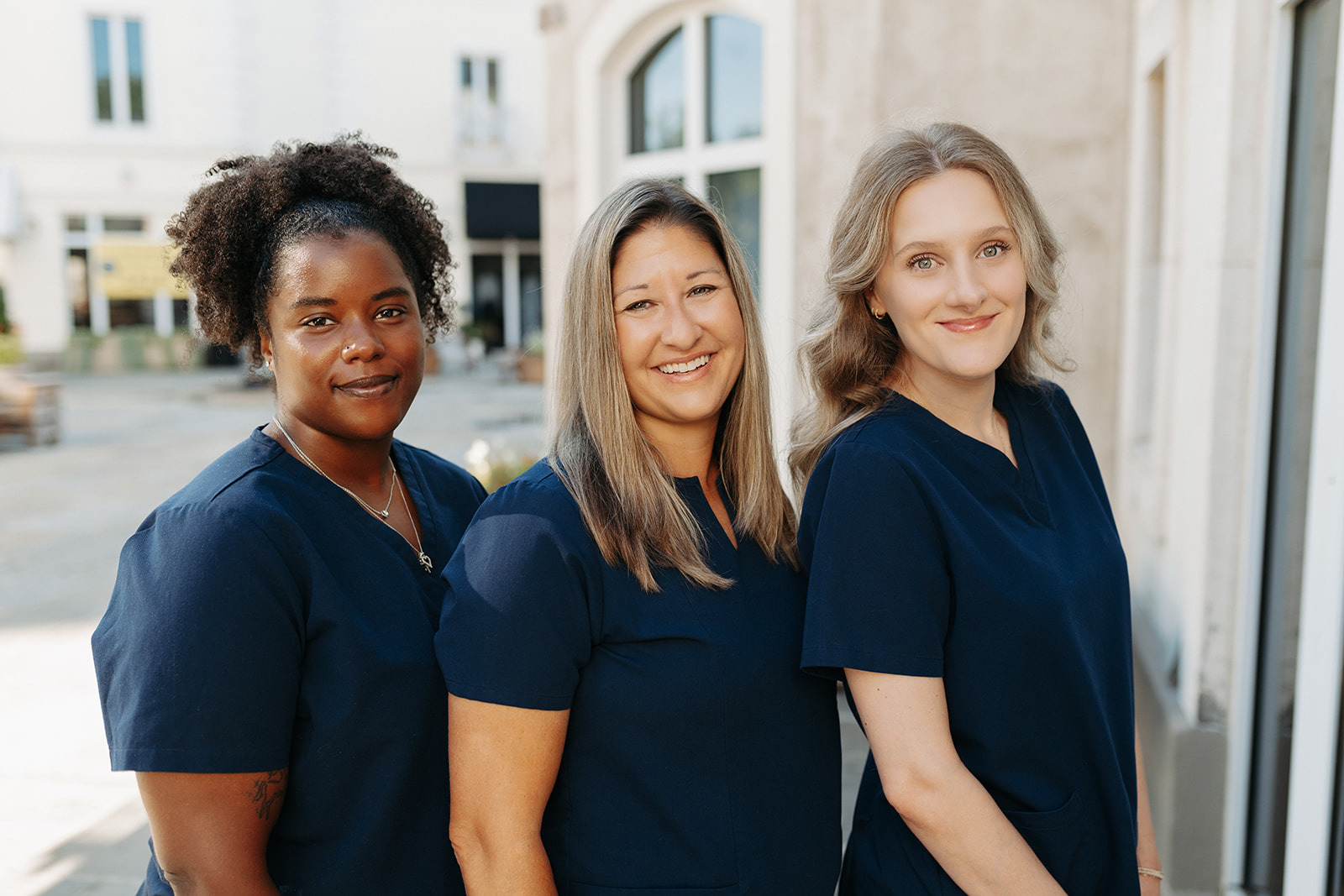 Three women in navy scrubs standing outdoors in front of a building, smiling, with a city street background.