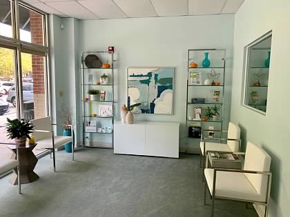Interior of a waiting area with chairs, shelves with decorative items, a painting, and a window with a view outside.