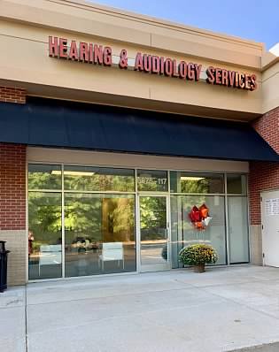 Storefront with sign reading 'Hearing & Audiology Services' above glass doors, a potted plant outside, and reflections of trees in the glass.