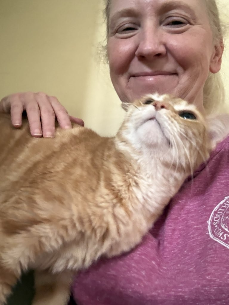 Person smiling with a ginger cat resting on their shoulder, close-up of face and cat's head, indoors.