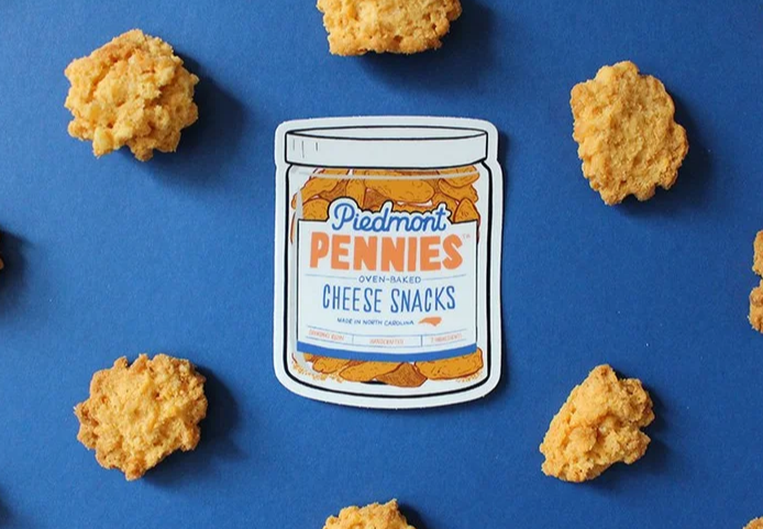 Jar of cheese snacks labeled 'Piedmont Pennies' surrounded by scattered cheese snack pieces on blue background.