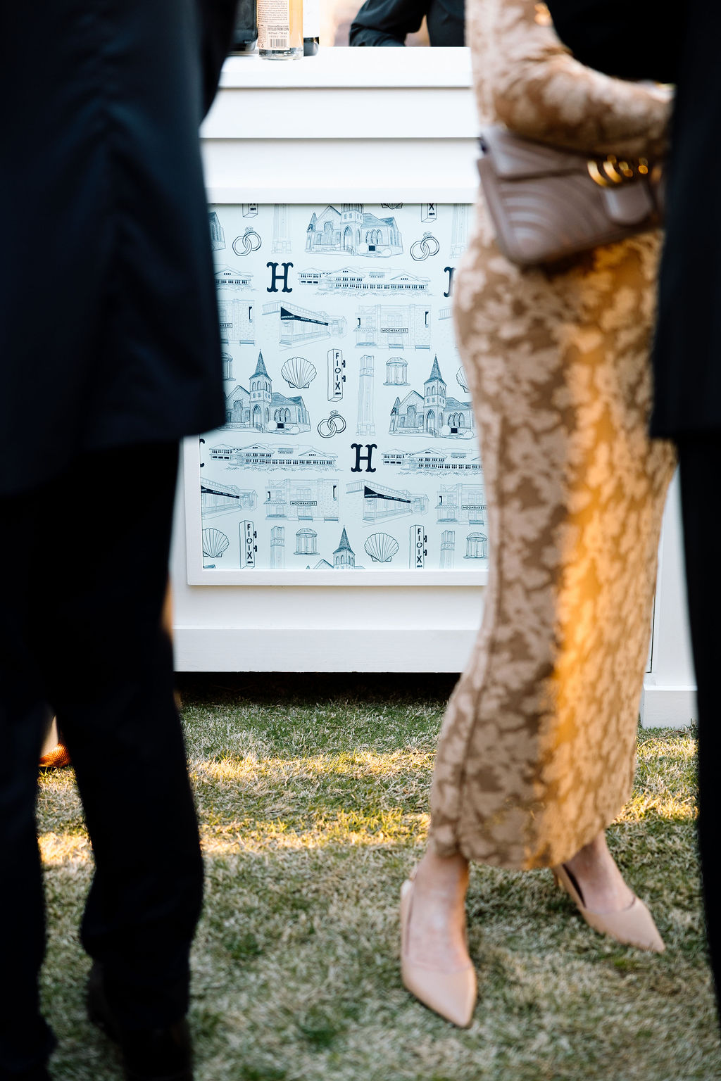 People standing outdoors near a decorated white display with illustrations, grass visible at the bottom, and two women in elegant clothing.