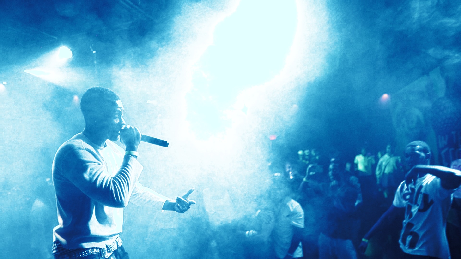 Person holding microphone performing on stage with a crowd in the background, blue lighting, and smoke effects.