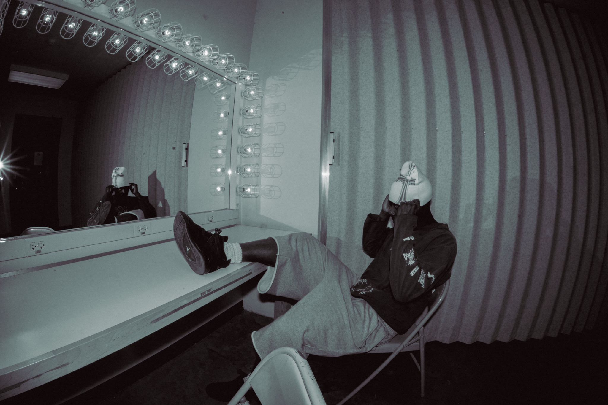 Person sitting with legs on a counter, drinking from a cup in a dressing room with striped curtains and mirror lights.