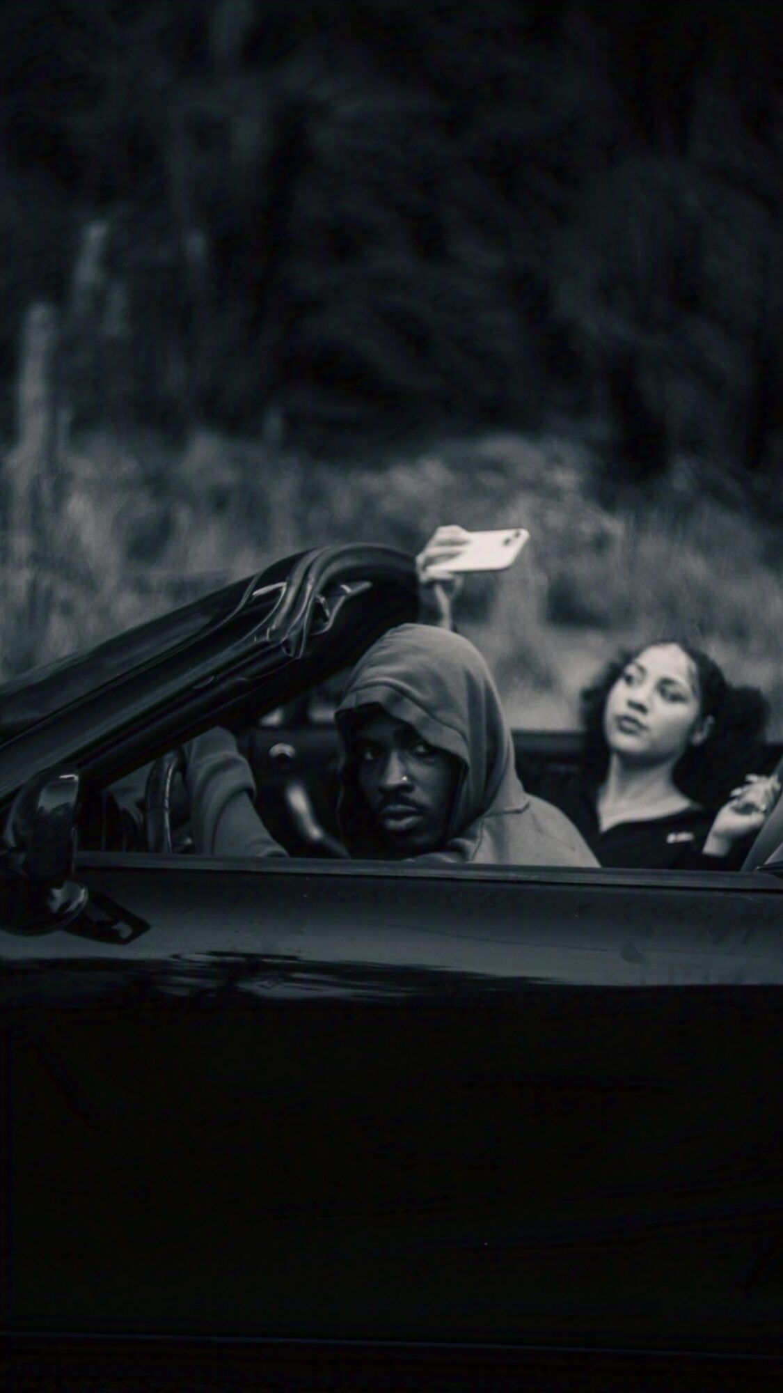 Two people in a car, one wearing a hoodie, the other with long hair, in a dark outdoor setting.