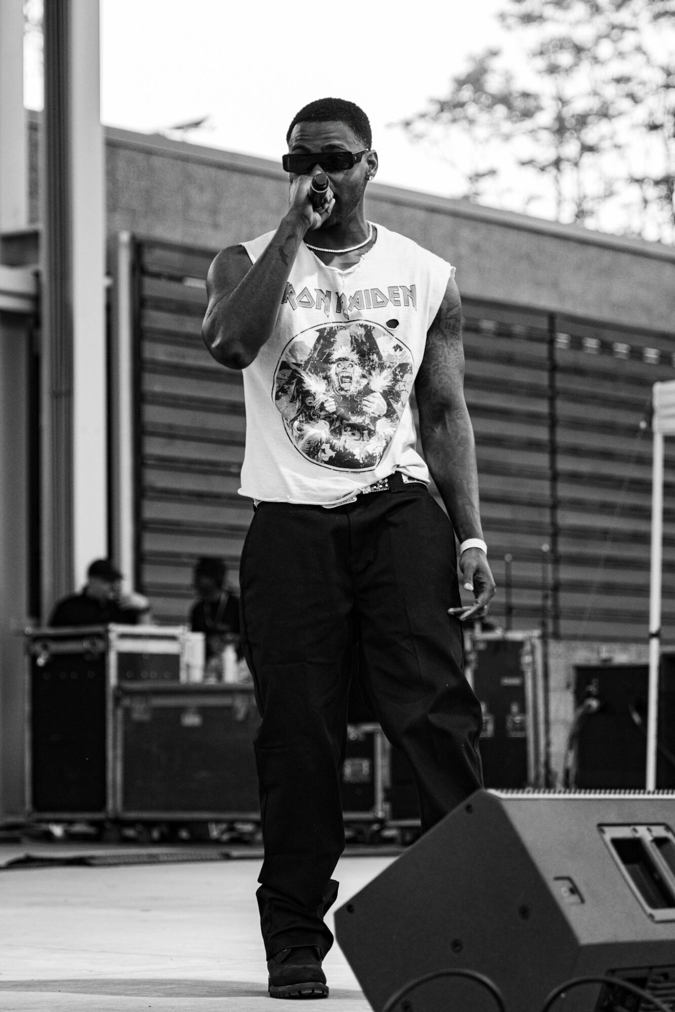 Man singing into microphone on stage outdoors, wearing sunglasses and a graphic t-shirt, with a fence and trees in background.