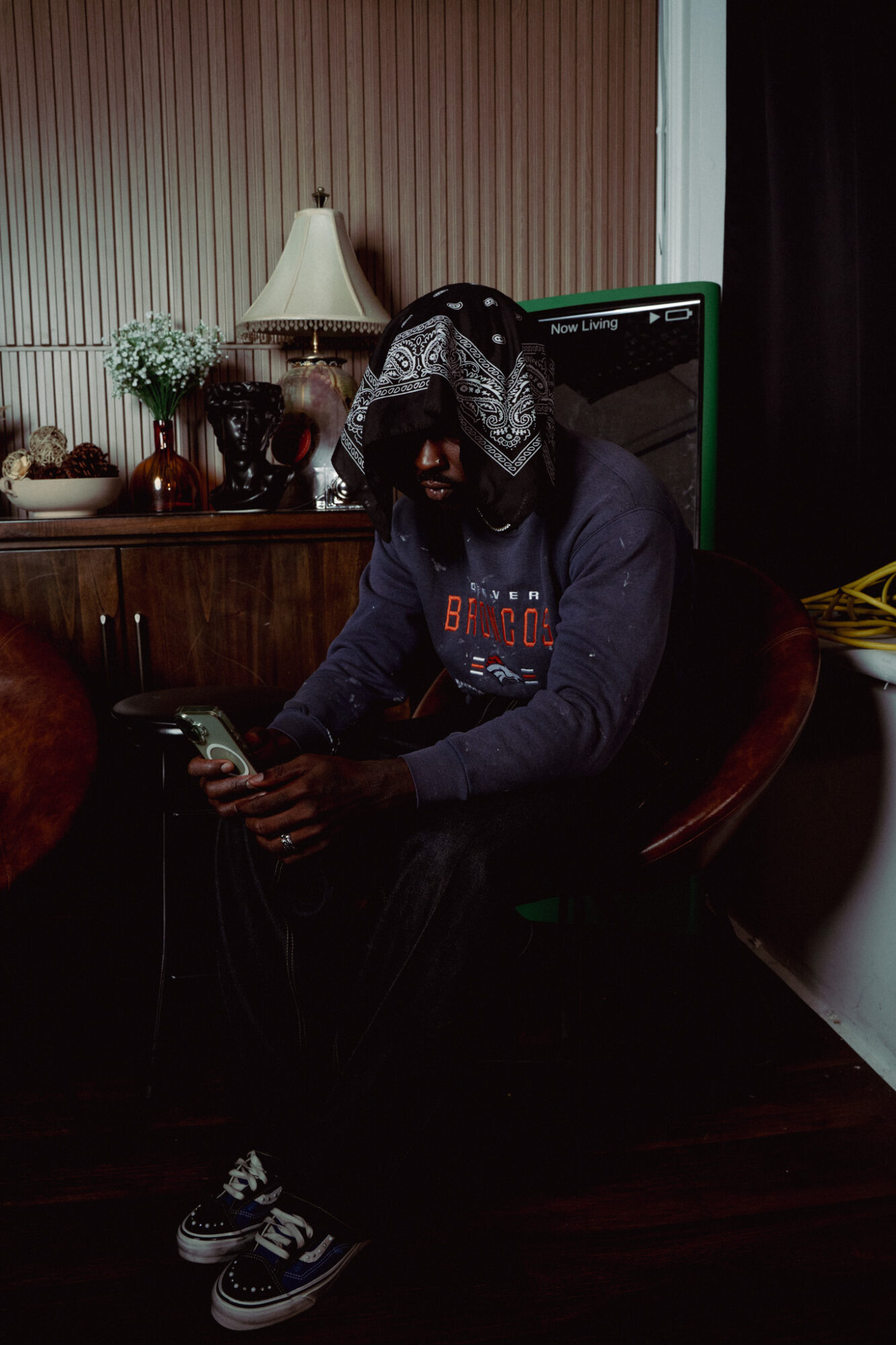 Person sitting in a room, wearing a hoodie and a bandana, looking at a phone, with a lamp and plants behind.