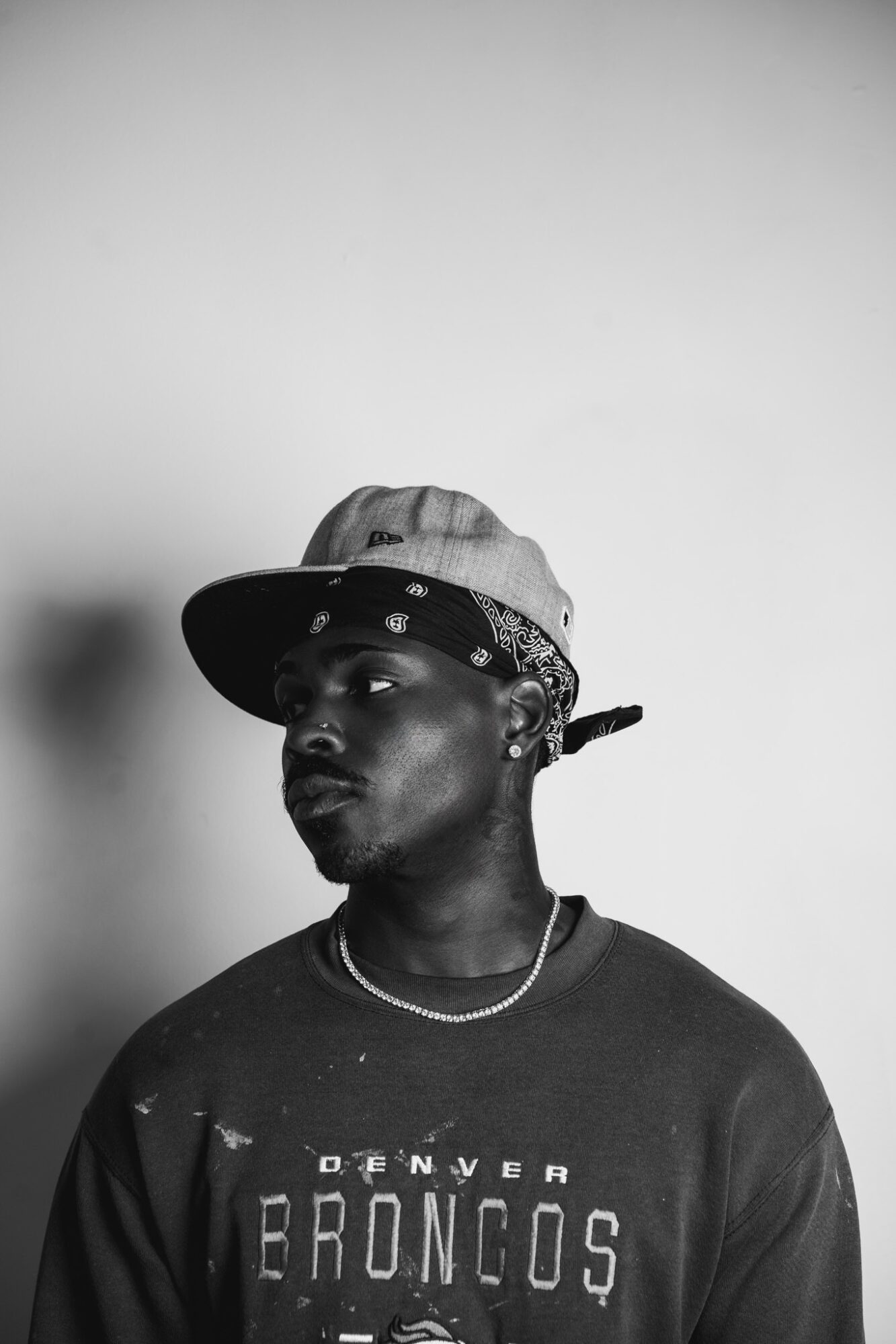 Young man wearing a cap, bandana, and a Denver Broncos shirt, looking to the side against a plain background.