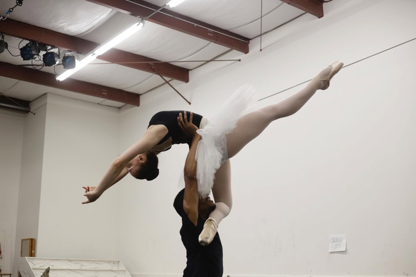 Ballet dancer in a white tutu being lifted horizontally by a partner in a dance studio with white walls and ceiling beams.