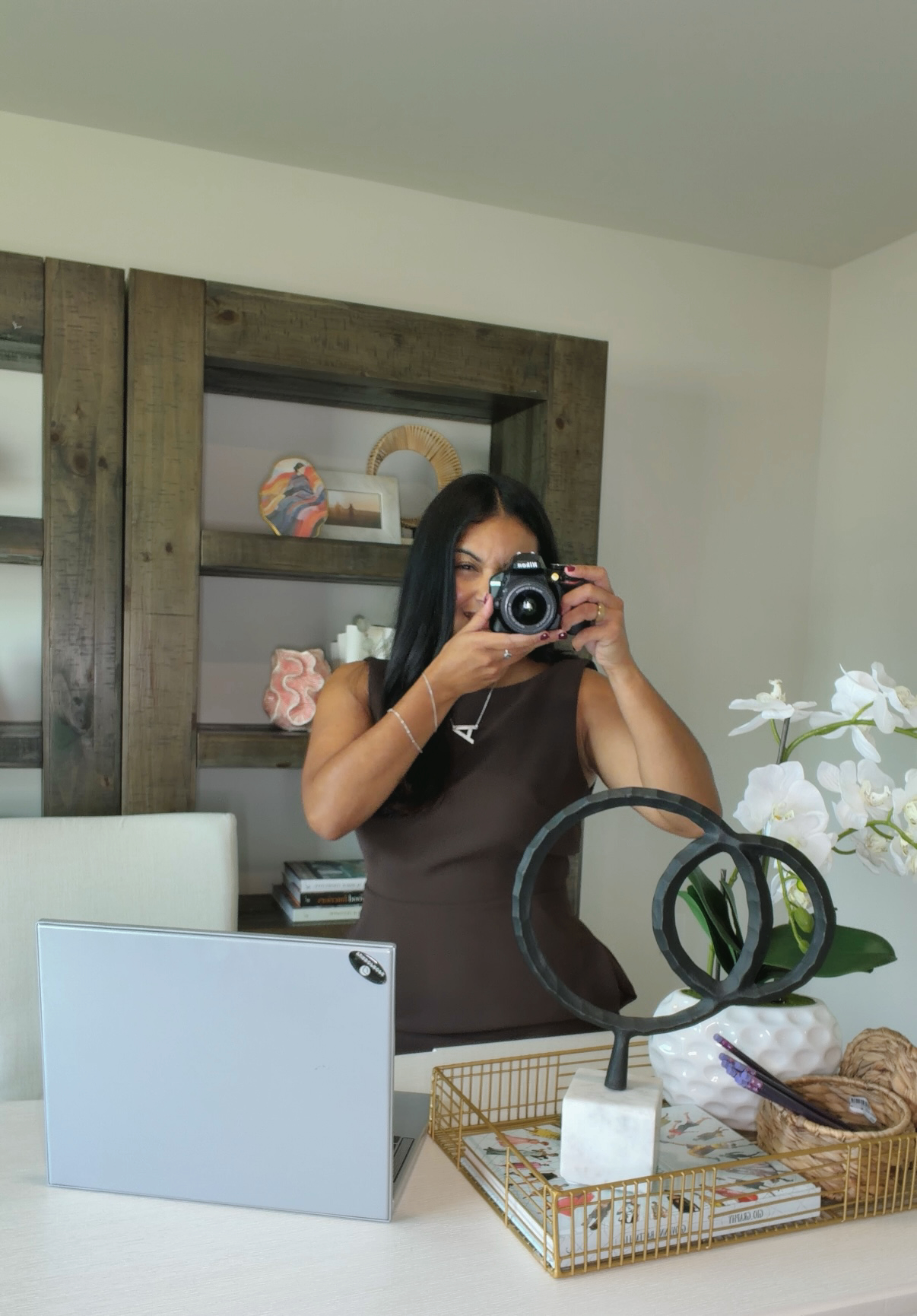 Person taking a photo in a mirror, with shelves and decorative items in the background, and a table with plants and objects in front.