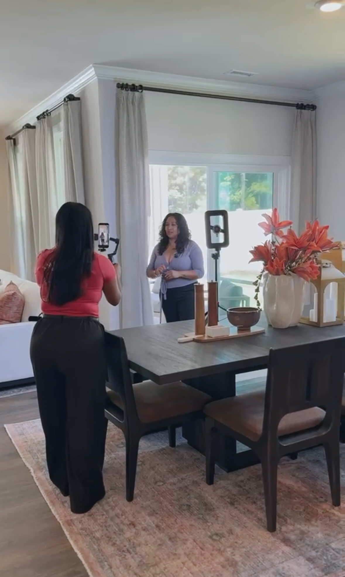 Two women stand in a bright living room, one taking a photo of the other near large windows with curtains, decorated with flowers.