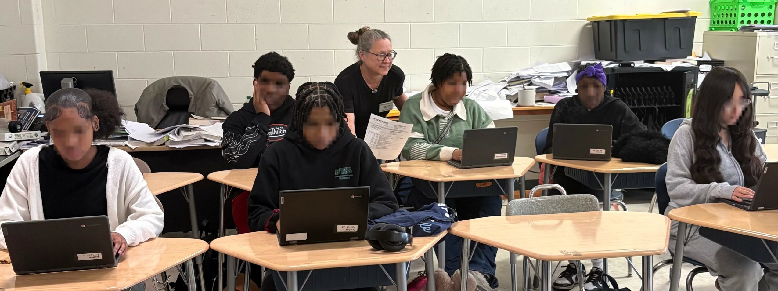 Group of students sitting at desks with laptops in a classroom, some looking at screens, others writing or talking.