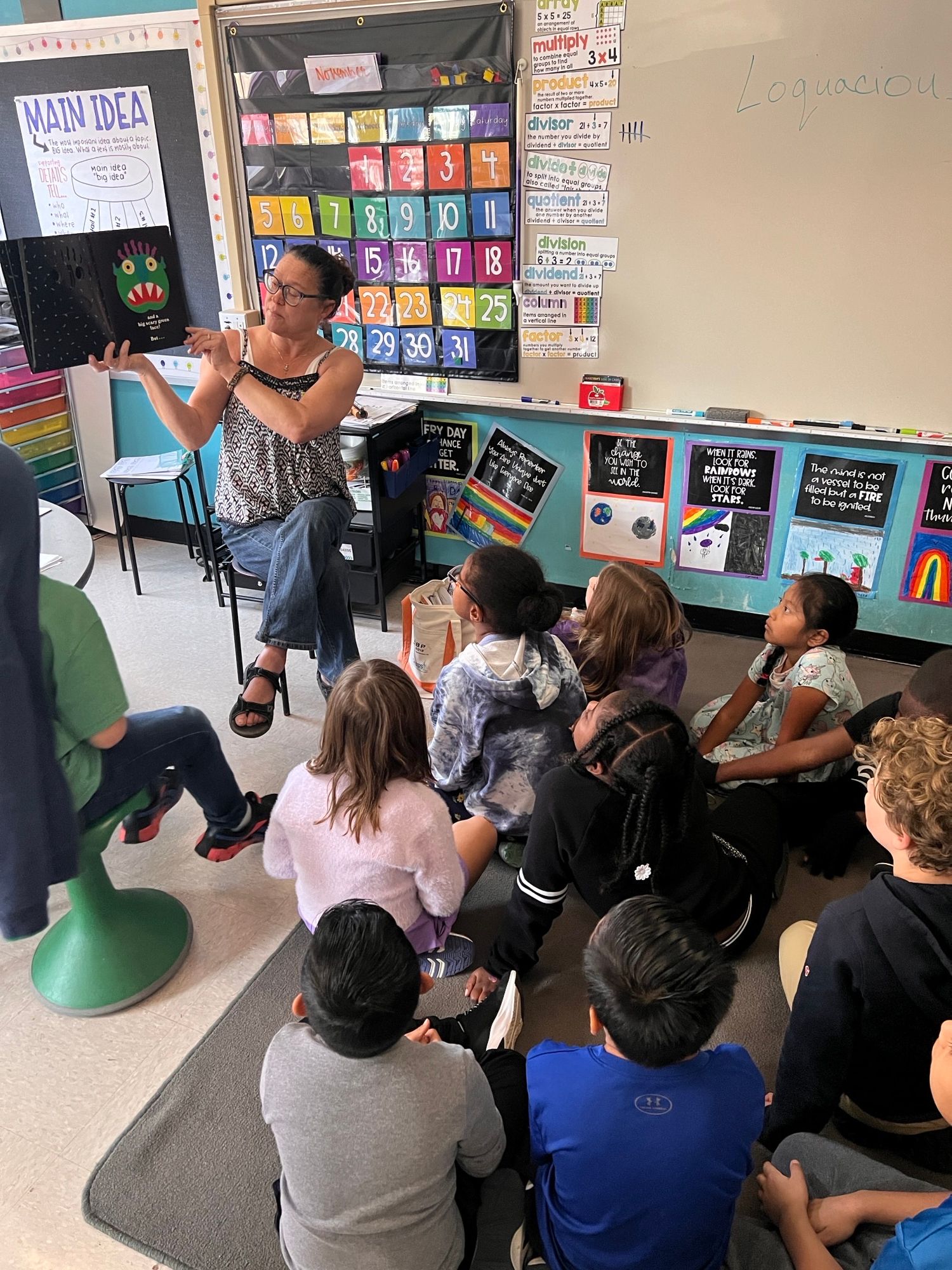 Group of people gathered around a woman speaking in a classroom with colorful educational posters and materials on the wall.
