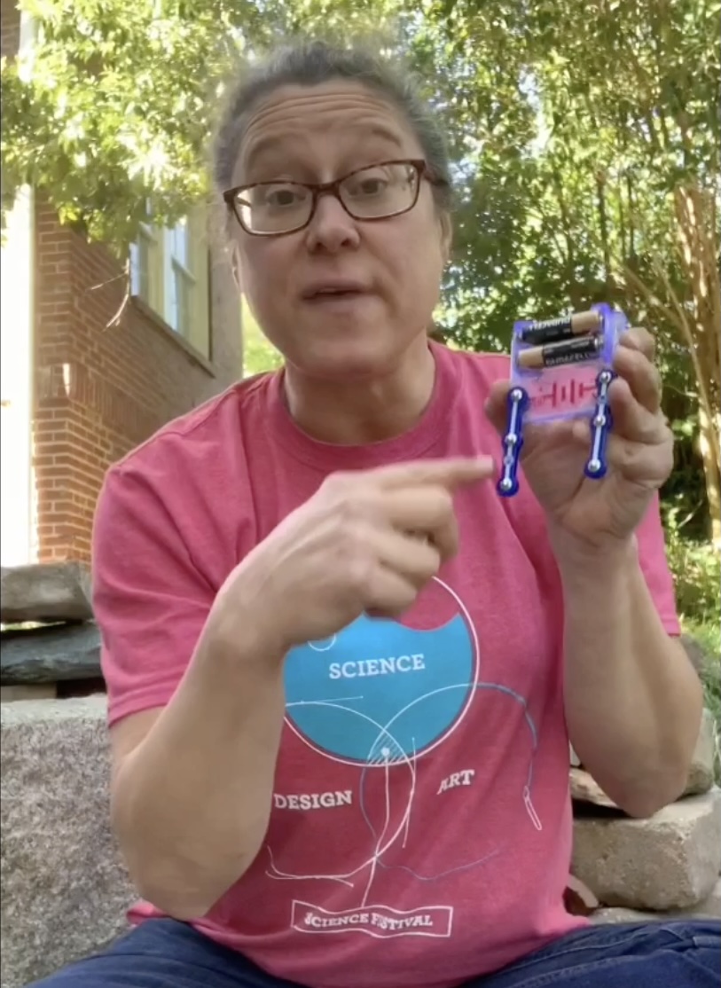 Woman with glasses holding a small robot, pointing at it outdoors with trees and a brick building in background.