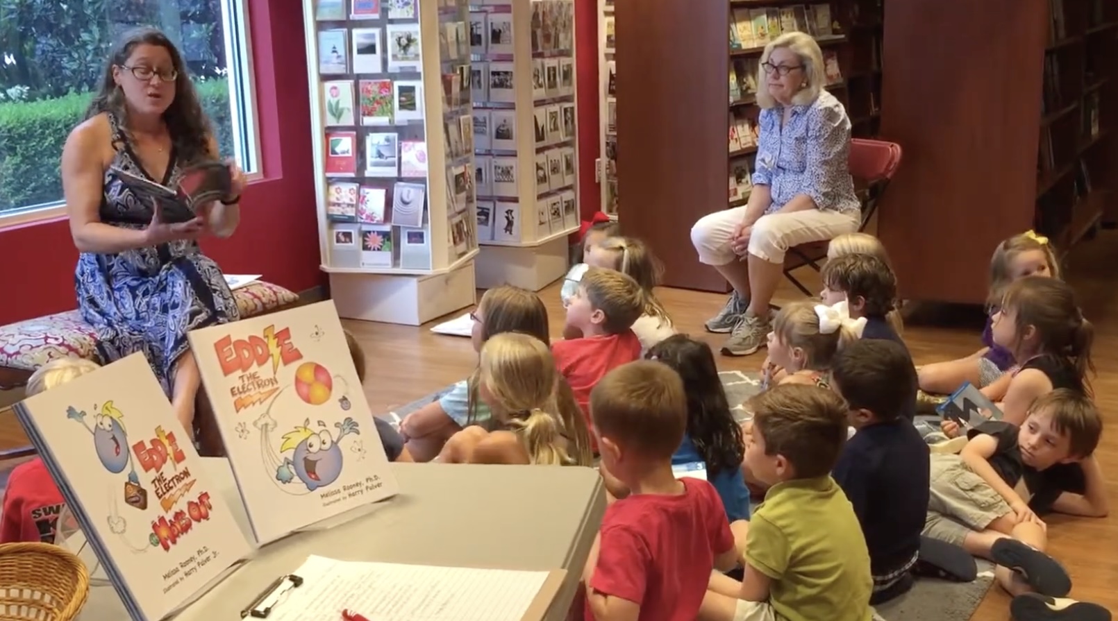 Children sitting on the floor listening to a woman reading a book in a library, with bookshelves in the background.