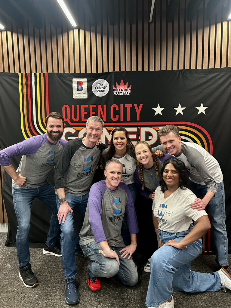 Group of eight people smiling and posing in front of a Queen City banner, some with arms around each other.