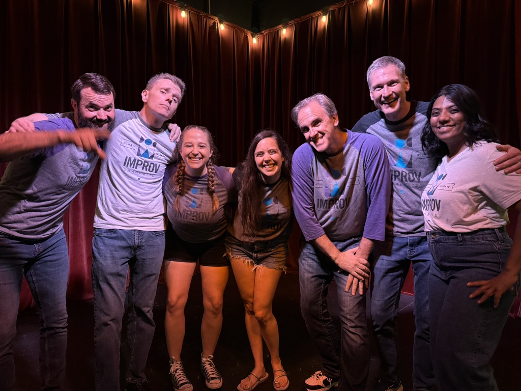 Group of seven people smiling and posing on stage with dark curtains and string lights behind them.