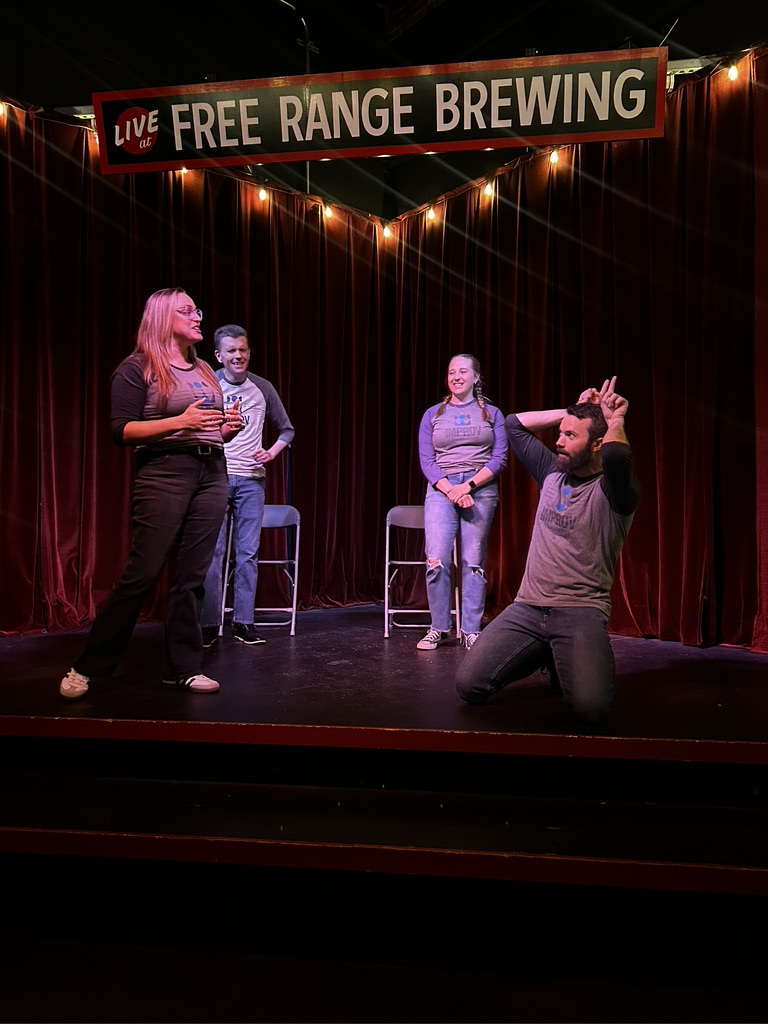 Four people on stage at a comedy event, with a banner reading 'Free Range Brewing' and string lights overhead.
