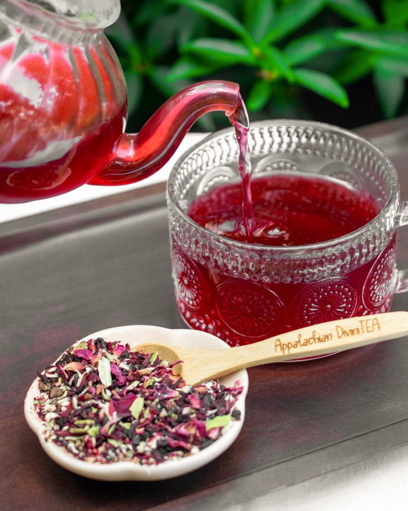 Red tea being poured into a glass cup with a bowl of dried tea leaves nearby, on a dark surface with green plants in background.