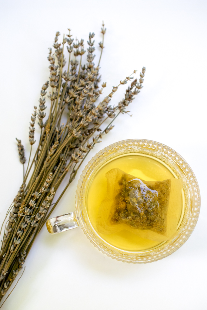 Dried lavender bunch next to a glass cup with herbal tea and a tea bag inside, on a white background.