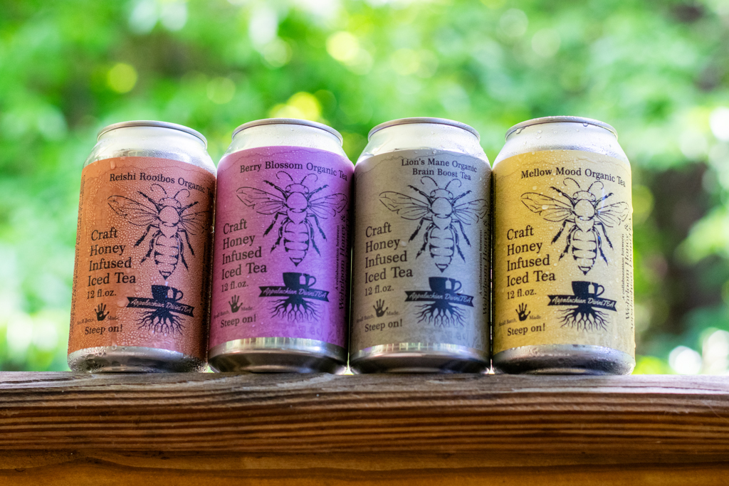 Four cans of craft honey-infused iced tea with colorful labels on a wooden surface, outdoors with green foliage background.